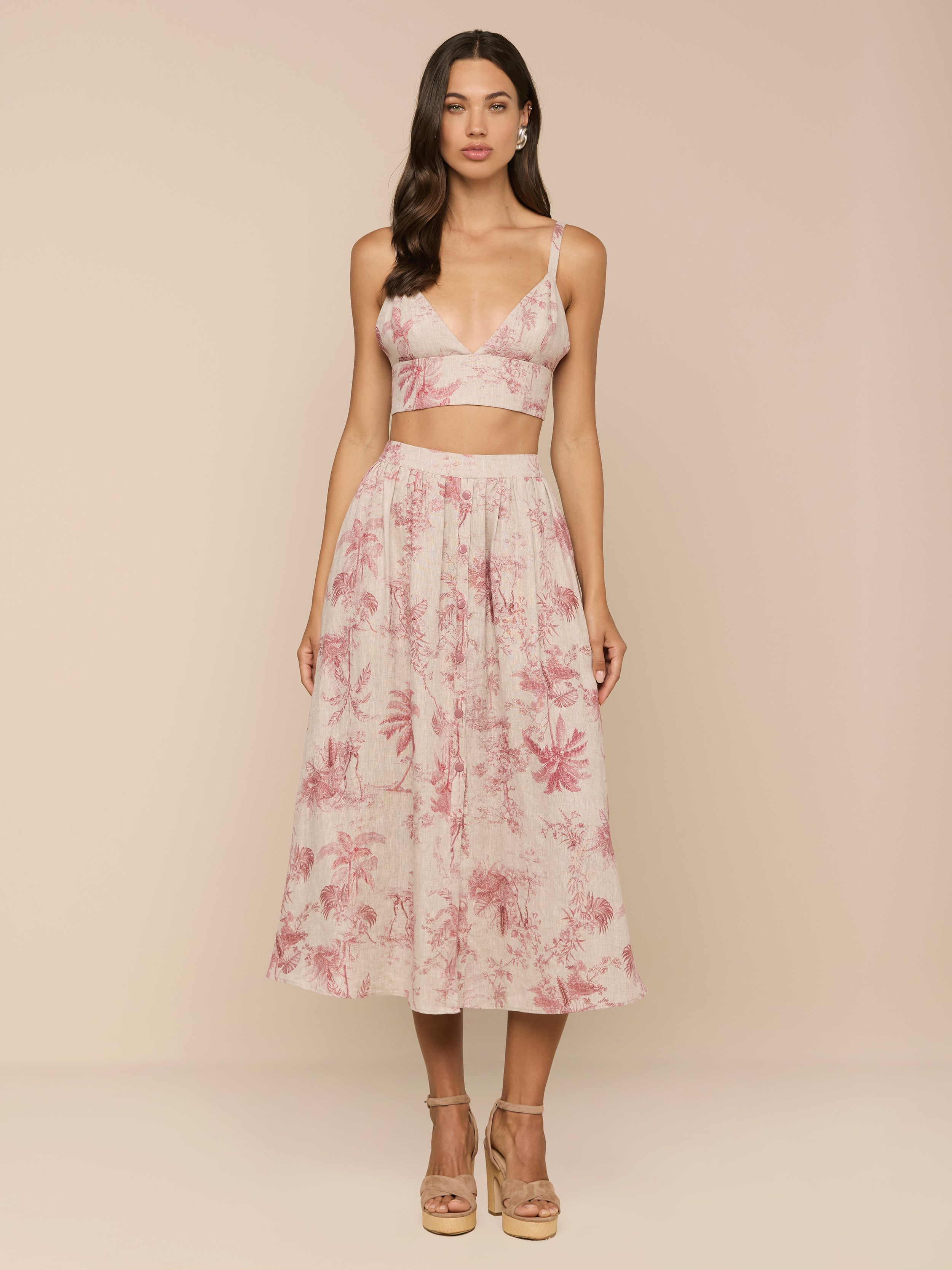 A woman stands against a beige background wearing the Seina Linen Midi Skirt, a high-waisted, flowy skirt with a pink floral pattern, paired with a sleeveless light crop top and beige platform heels.