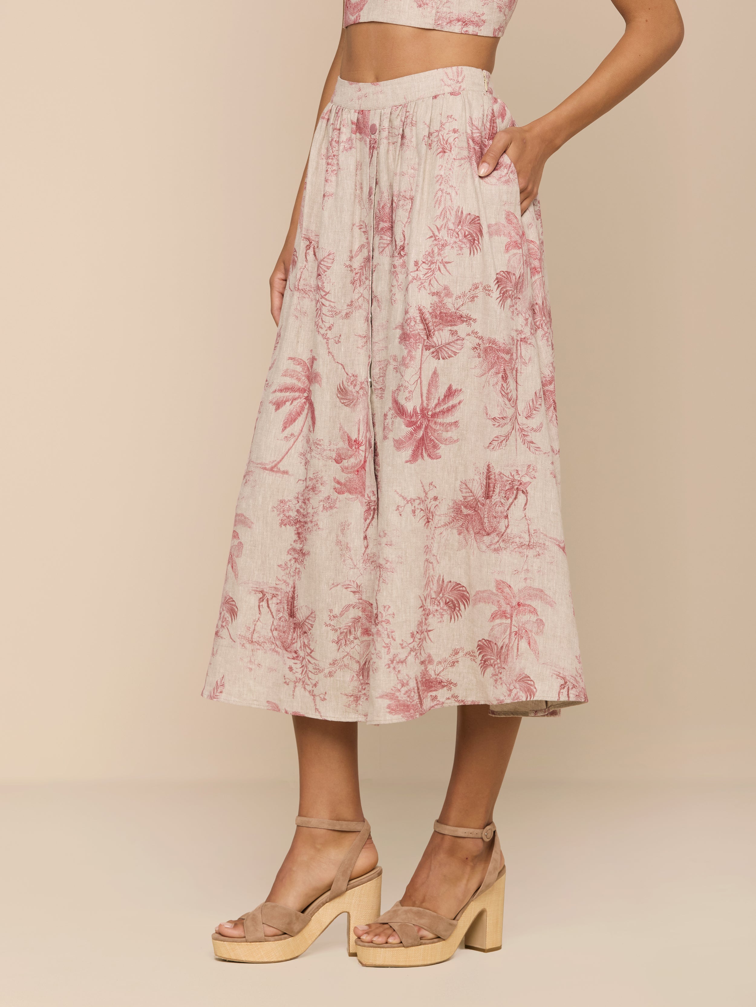 A woman wearing the Seina Linen Midi Skirt, featuring beige pleats with pink floral prints, a matching crop top, and tan platform sandals stands against a plain beige background.