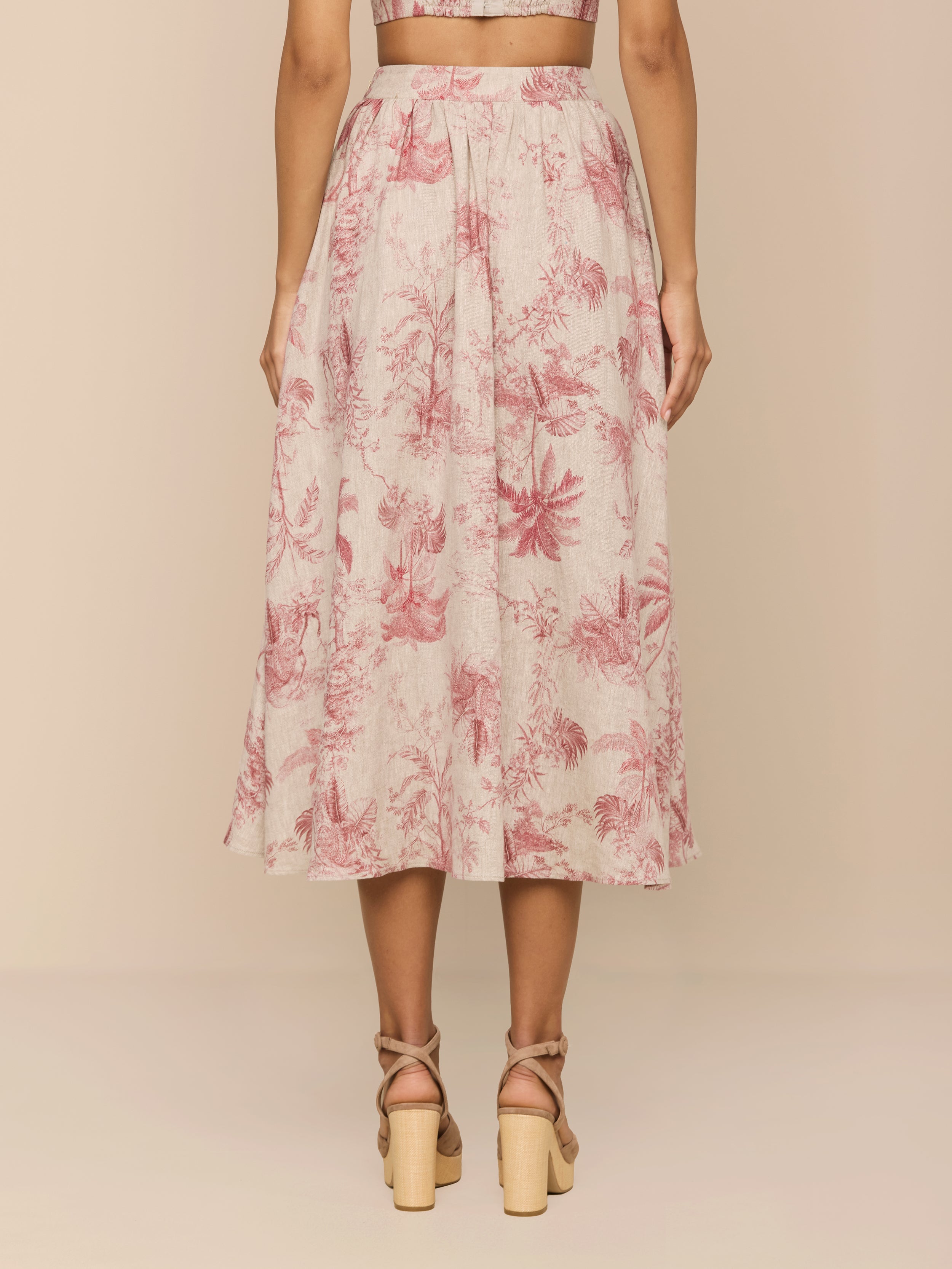 A woman stands with her back to the camera wearing the Seina Linen Midi Skirt, featuring a high waist and pink and white jungle toile print, paired with nude strappy block-heeled sandals.