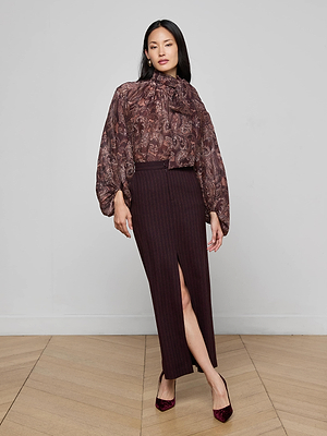 A woman poses indoors in a sheer paisley blouse with voluminous sleeves and a high neck, paired with the Maceline Pinstriped Maxi Skirt featuring a front slit and burgundy heels. The background is plain and minimal.