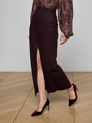 Someone wearing the Maceline Pinstriped Maxi Skirt with a front slit, a patterned billowy-sleeve blouse, and burgundy velvet heels stands on a wooden floor.