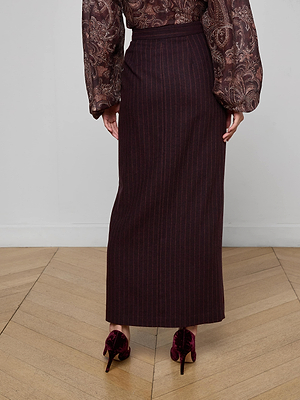 A person stands on a wood floor, seen from behind, wearing the Maceline Pinstriped Maxi Skirt with a brown patterned billowy-sleeved blouse and burgundy velvet high-heeled shoes.