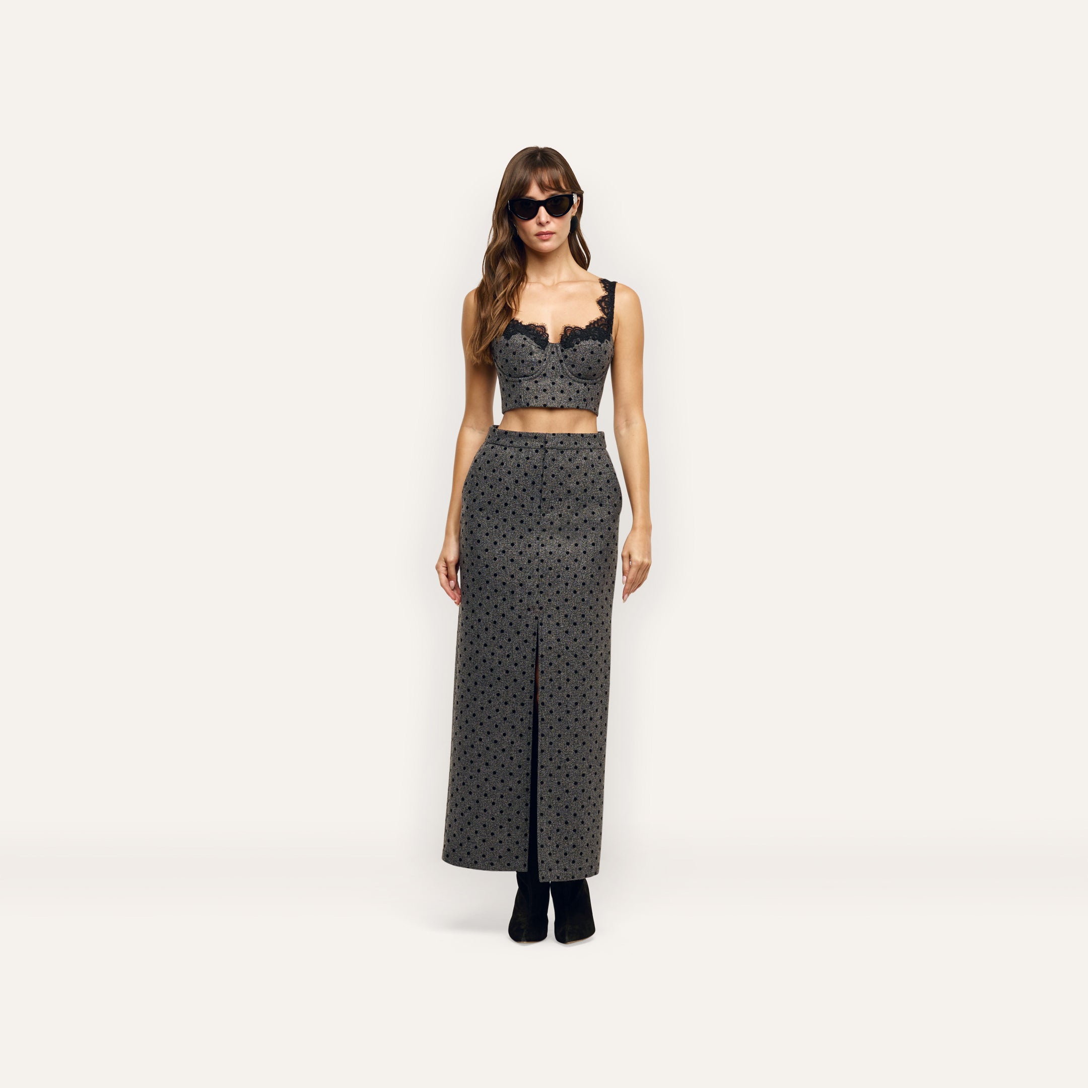 A model wearing a grey, velvet polka-dot Maceline Maxi Skirt with a front slit and a matching cropped bustier top with black lace trim.