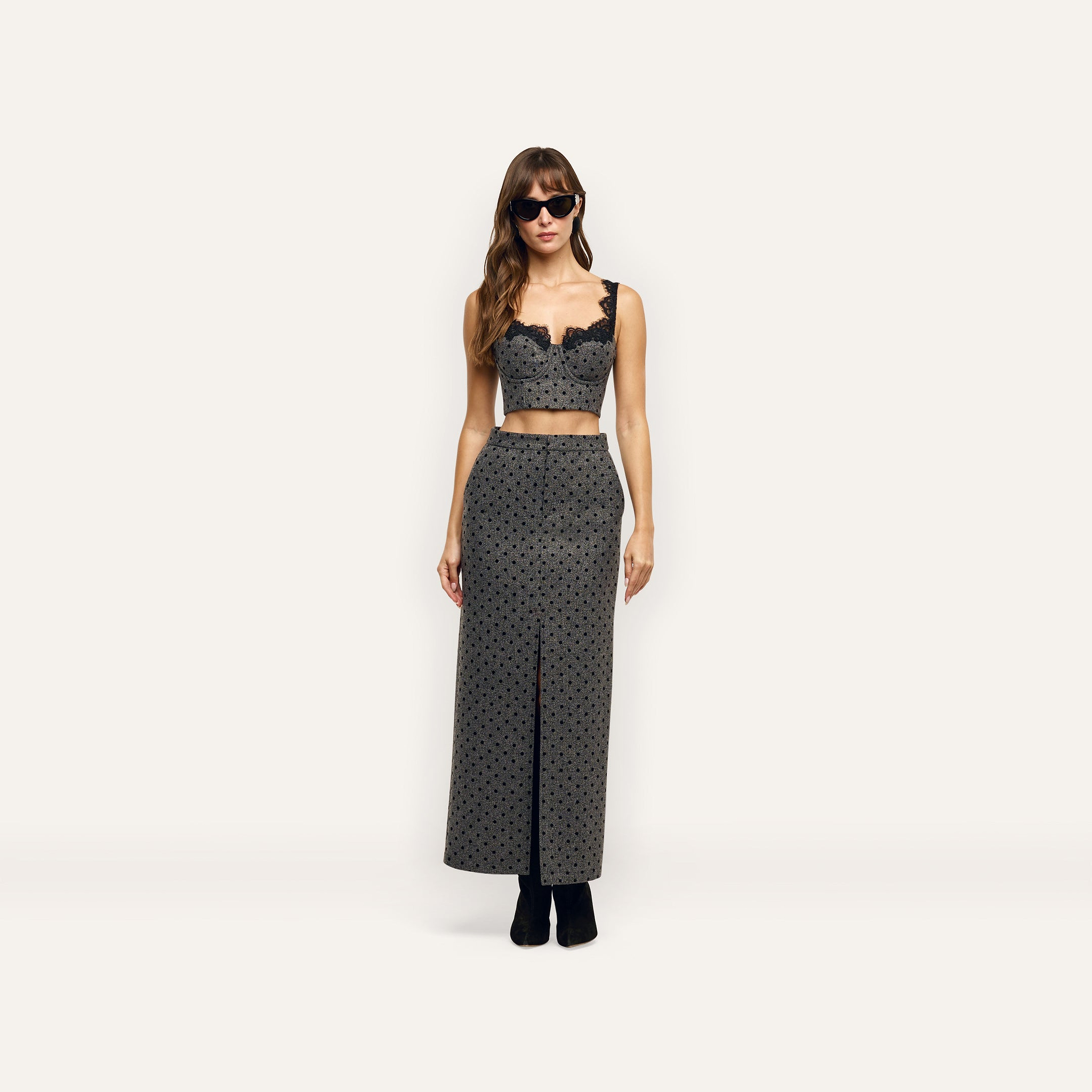 A model wearing a grey, velvet polka-dot Maceline Maxi Skirt with a front slit and a matching cropped bustier top with black lace trim.
