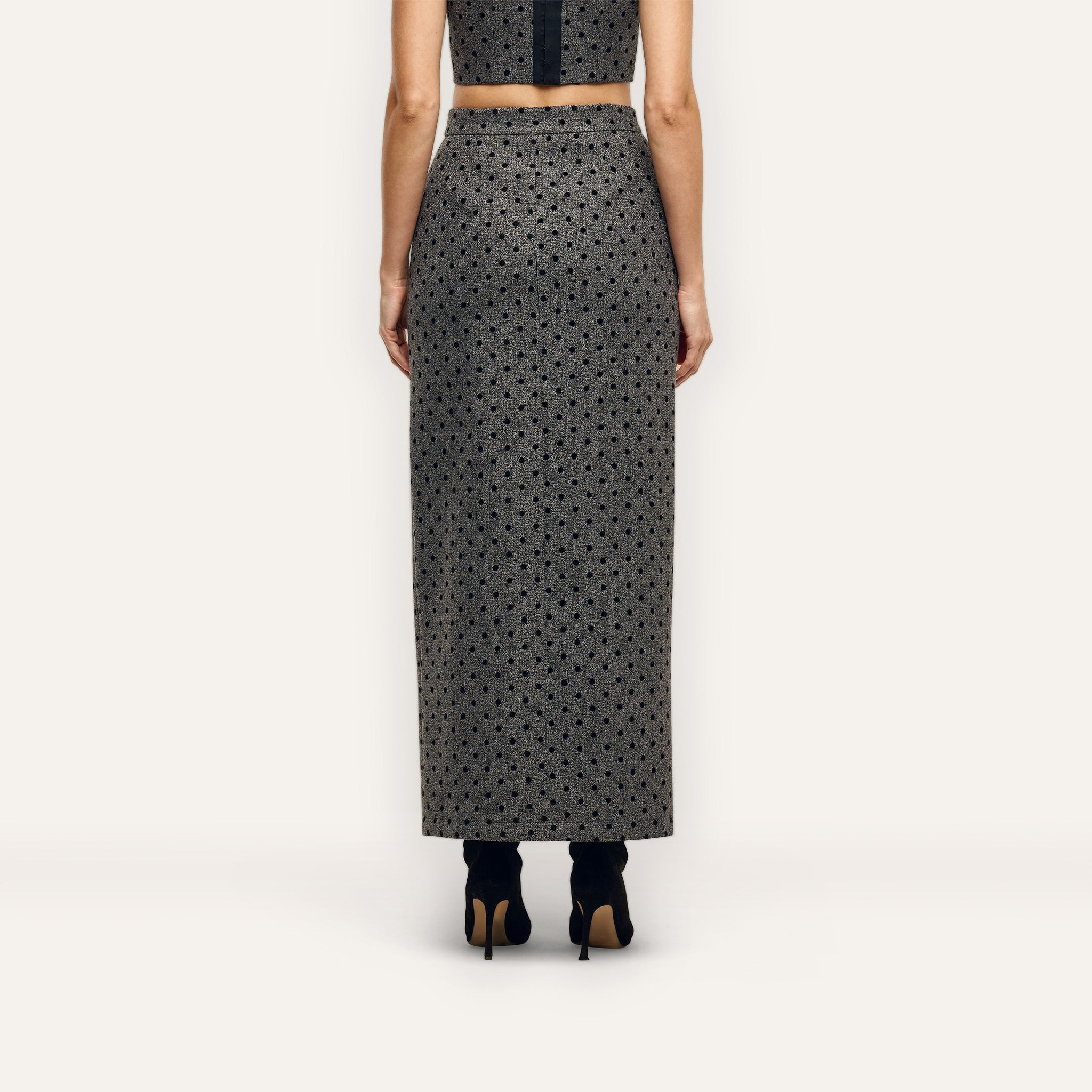 A person viewed from behind, wearing a grey polka-dot Maceline Maxi Skirt and black heeled boots.