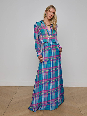 A woman with blonde hair stands on a wooden floor, wearing the Patricia Plaid Maxi Skirt by L'AGENCE Sale in vibrant blue, pink, and purple. She completes her look with layered necklaces and earrings.