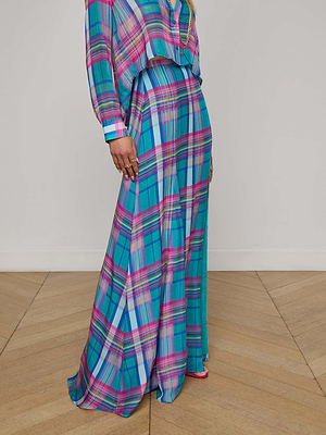 A person stands on a light wood herringbone floor against a plain white wall, wearing the L'AGENCE Sale Patricia Plaid Maxi Skirt paired with a vibrant blue, pink, and purple plaid viscose chiffon blouse.