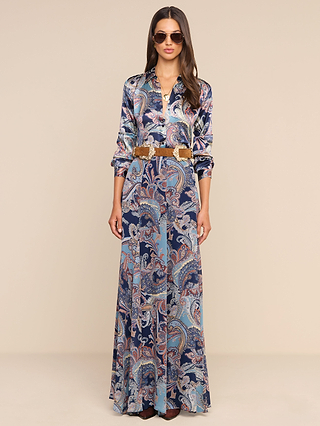 A woman stands against a plain beige background, wearing the flowing Patrica Maxi Skirt in elegant midnight blue with a paisley print, paired with a wide brown belt and sunglasses for a chic look.