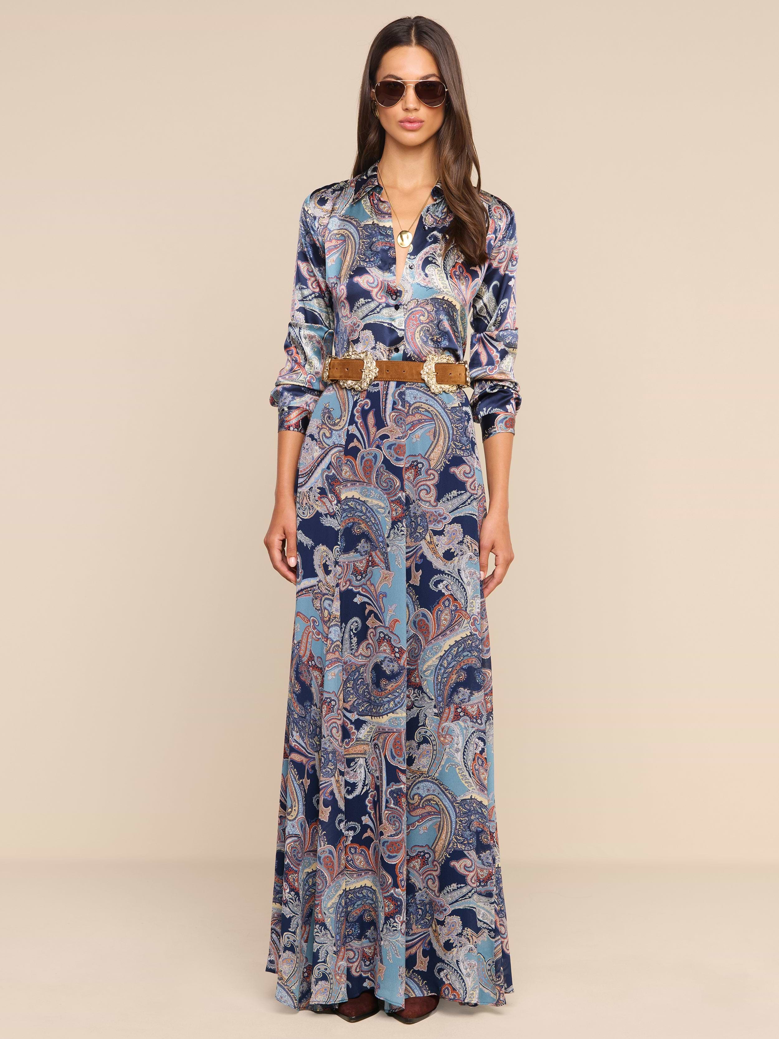 A woman wearing sunglasses and a long-sleeved top paired with the Patrica Maxi Skirt, featuring a floor-length paisley print and a brown belt, stands against a plain white background.