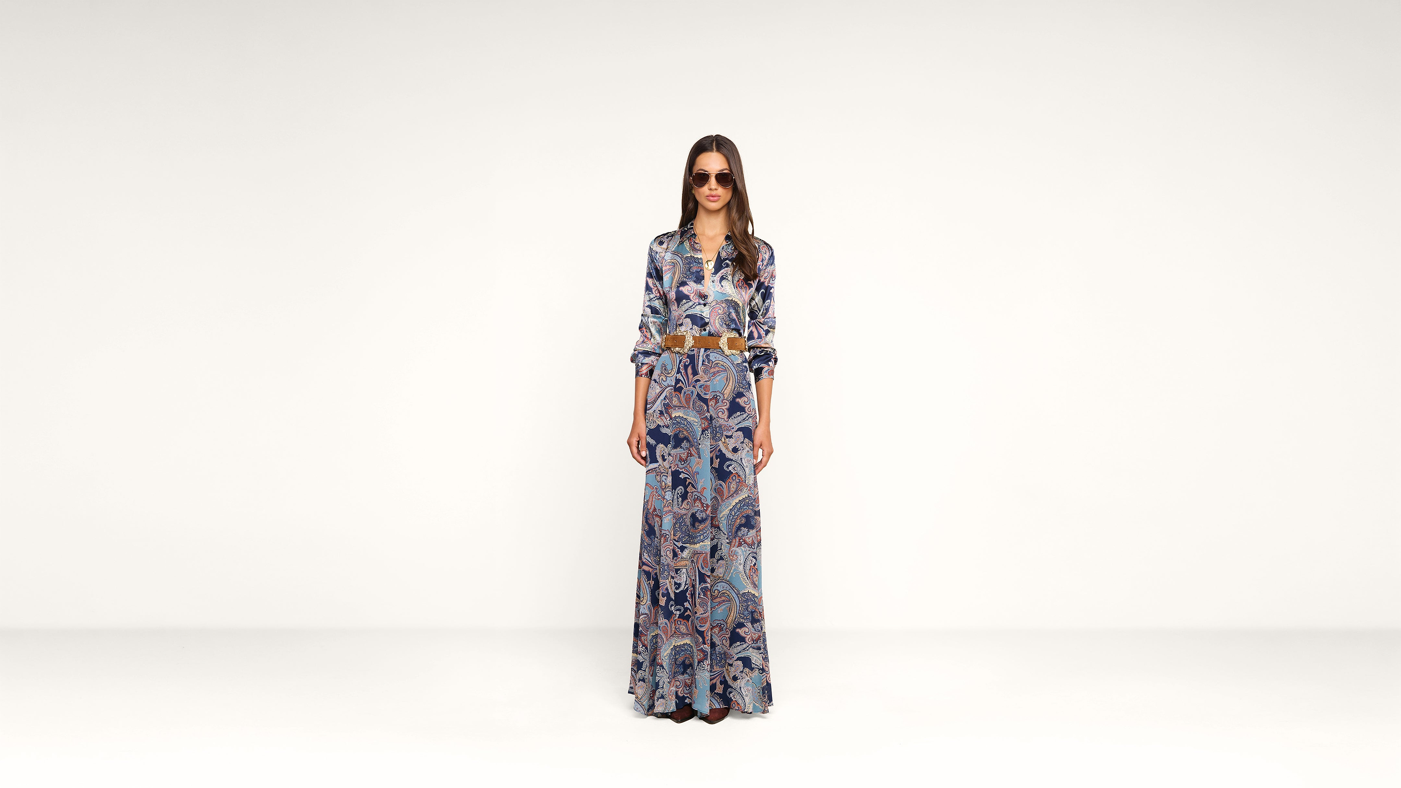 A woman wearing sunglasses and a long, colorful patterned dress stands against a plain white background, looking straight at the camera with her hands by her sides.