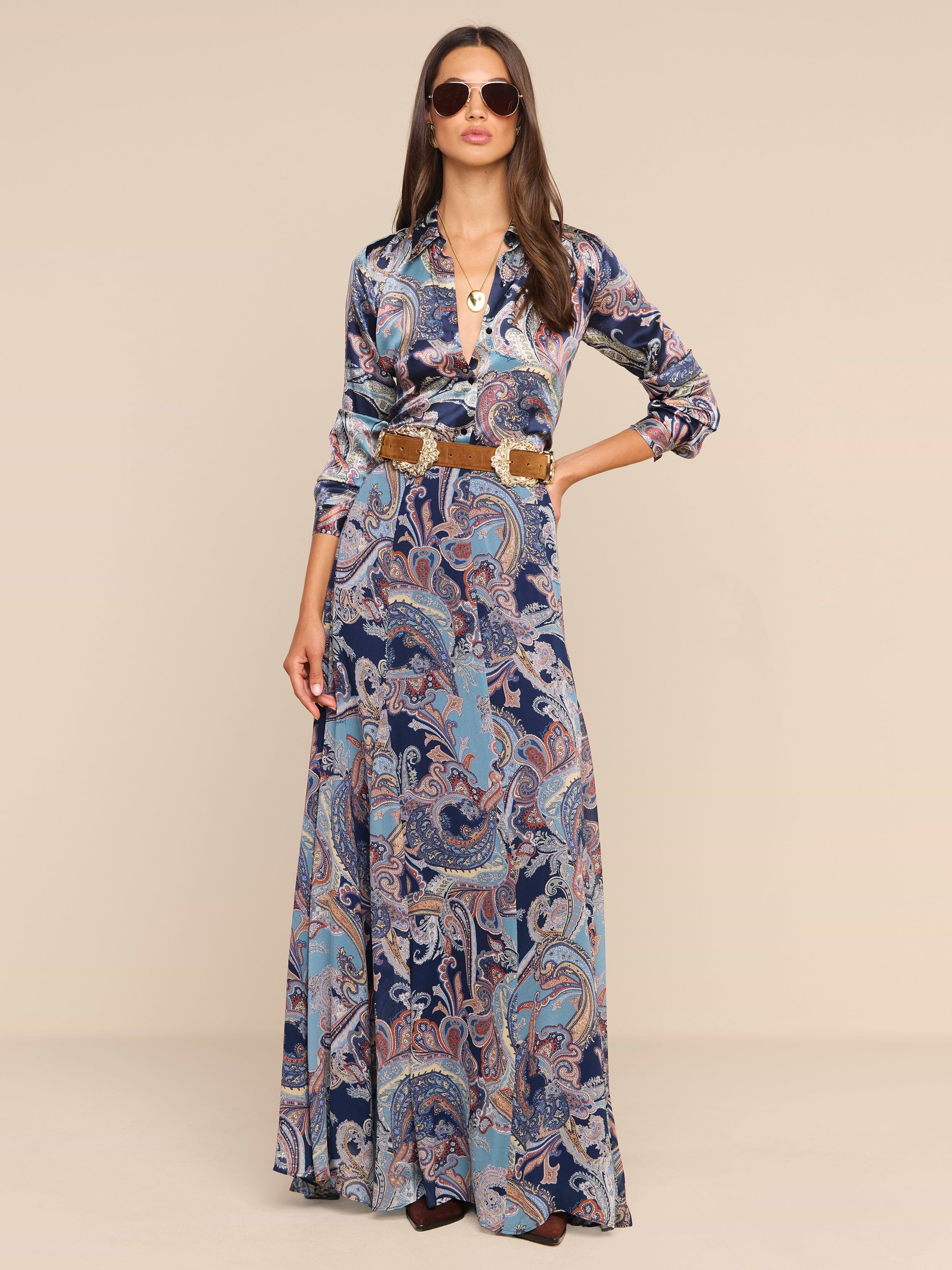 A woman wearing sunglasses, the Patrica Maxi Skirt in a long paisley print midnight blue design, and a brown belt stands confidently against a plain white background.