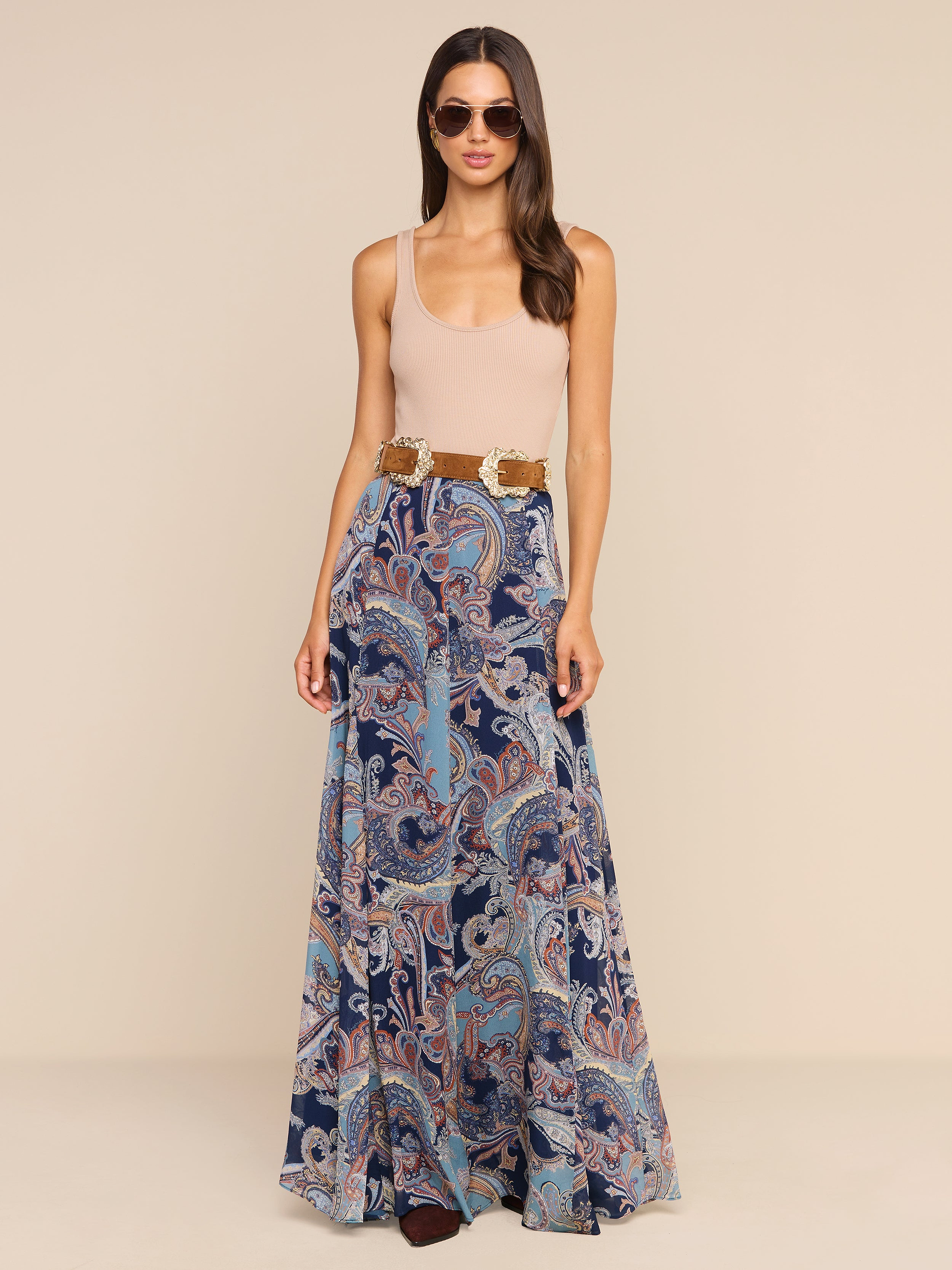 A woman wearing sunglasses, a beige sleeveless top, and the Patrica Maxi Skirt in midnight blue featuring a flowing paisley print with a wide belt, stands against a plain white background.