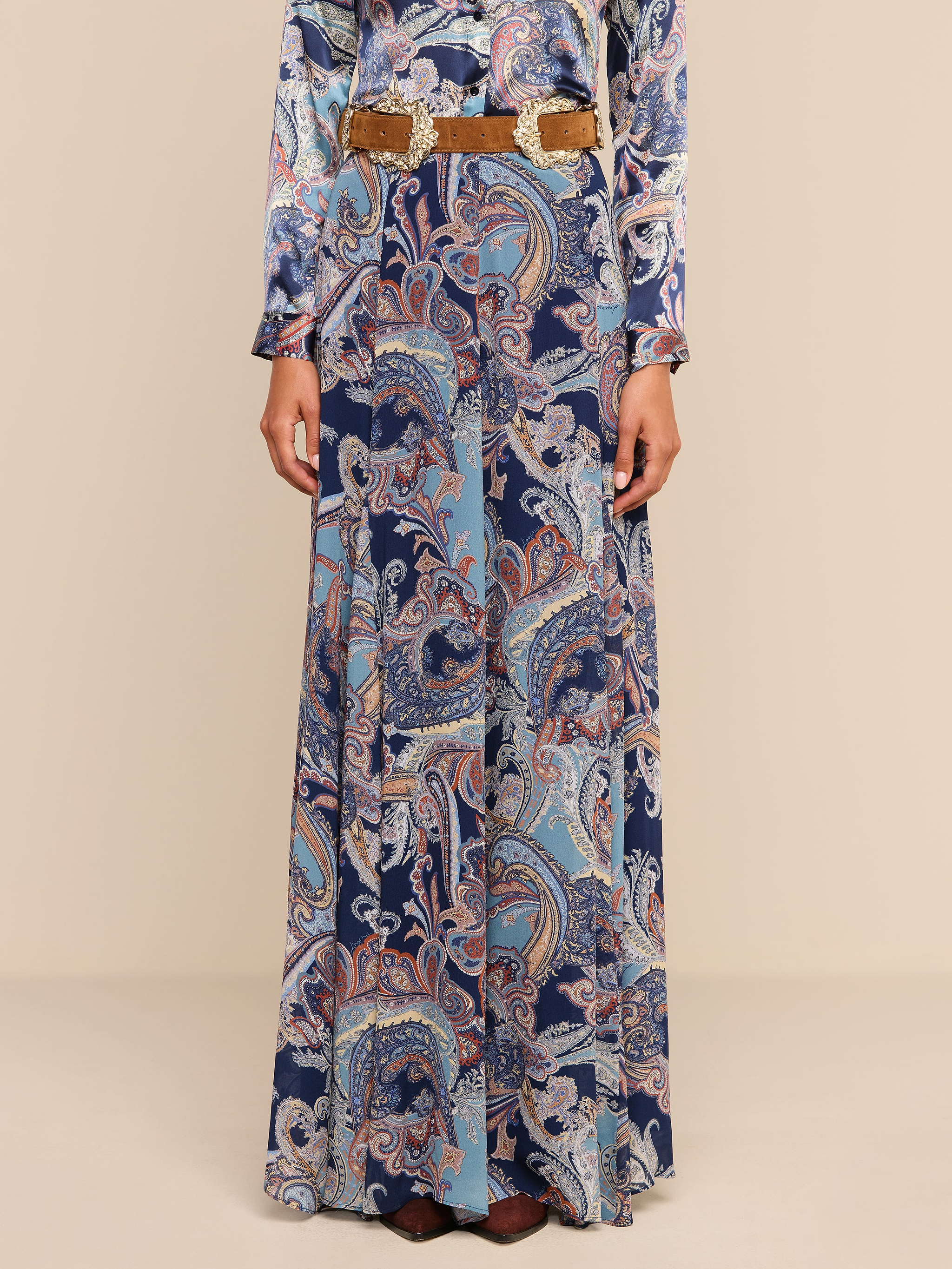 A person wearing the Patrica Maxi Skirt in a midnight blue paisley print with a tan belt, standing against a plain white background. Only the body below the shoulders is visible.