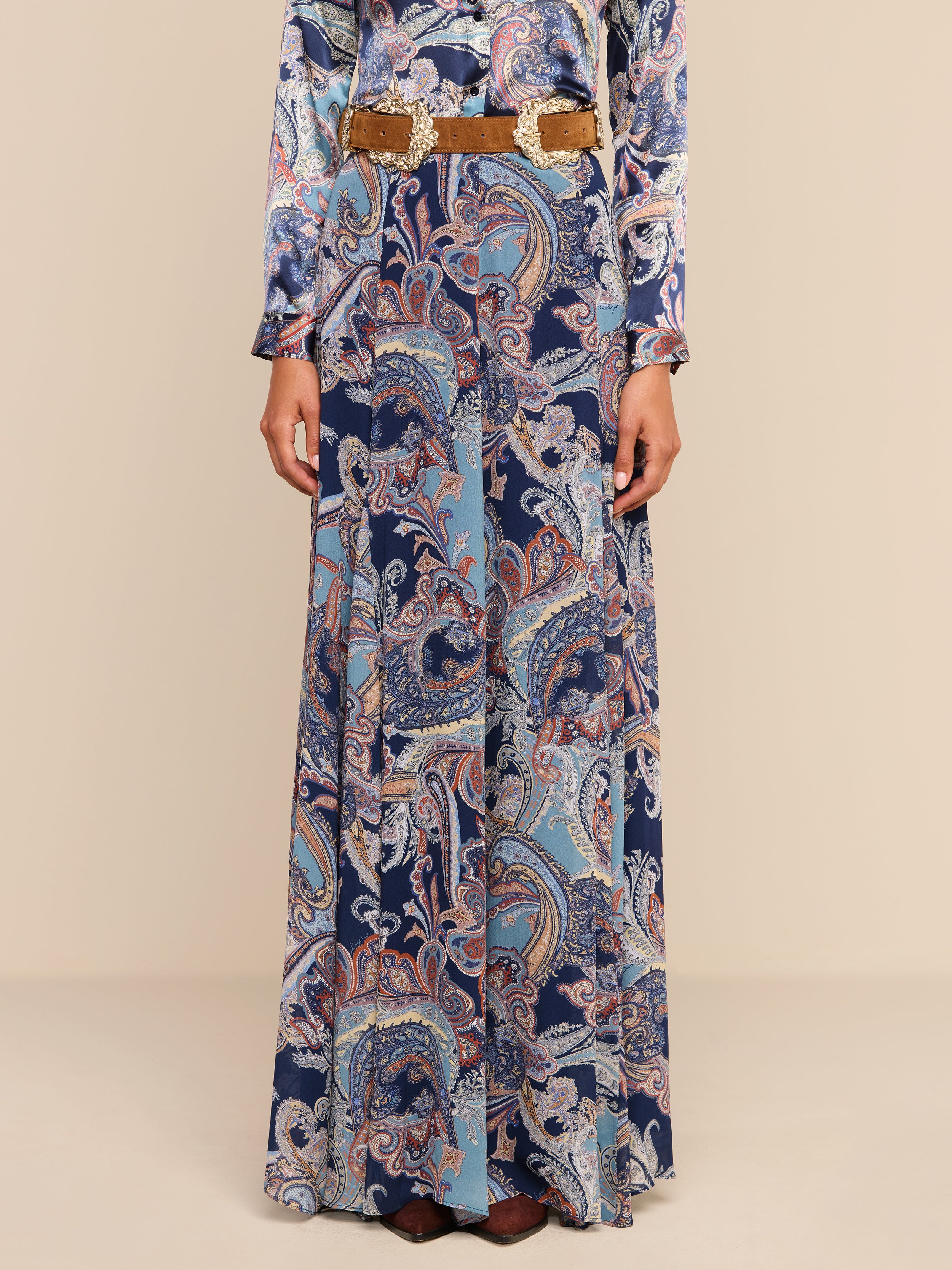 A person wearing the Patrica Maxi Skirt in a midnight blue paisley print with a tan belt, standing against a plain white background. Only the body below the shoulders is visible.