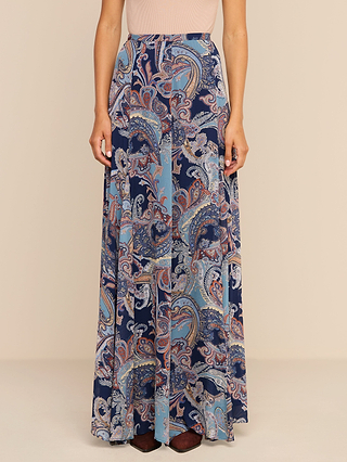 Wearing the Patrica Maxi Skirt—a wide-leg maxi silhouette adorned with a vibrant paisley print in blue, white, and peach—a person stands against a plain background, paired with a light pink sleeveless top.