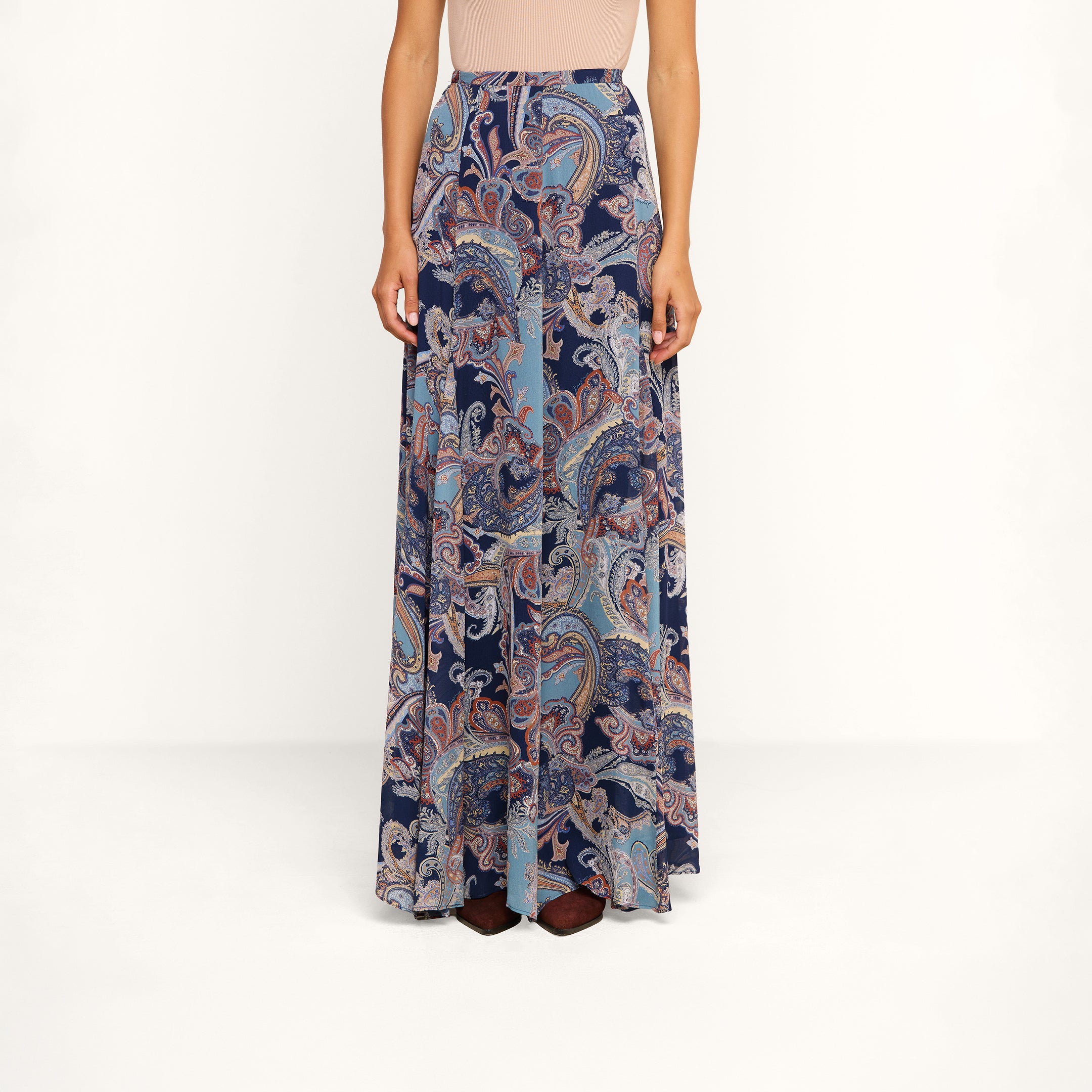 Wearing the Patrica Maxi Skirt, a long, flowy maxi skirt featuring a blue and pink paisley print, and a sleeveless beige top, a person stands against a plain background.