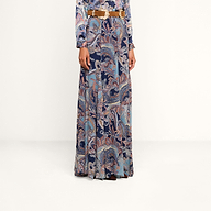 A person is wearing the Patrica Maxi Skirt, a floor-length paisley print skirt in shades of blue, purple, and pink, paired with a long-sleeved top and cinched at the waist with a brown belt. Their head is not visible in the image.