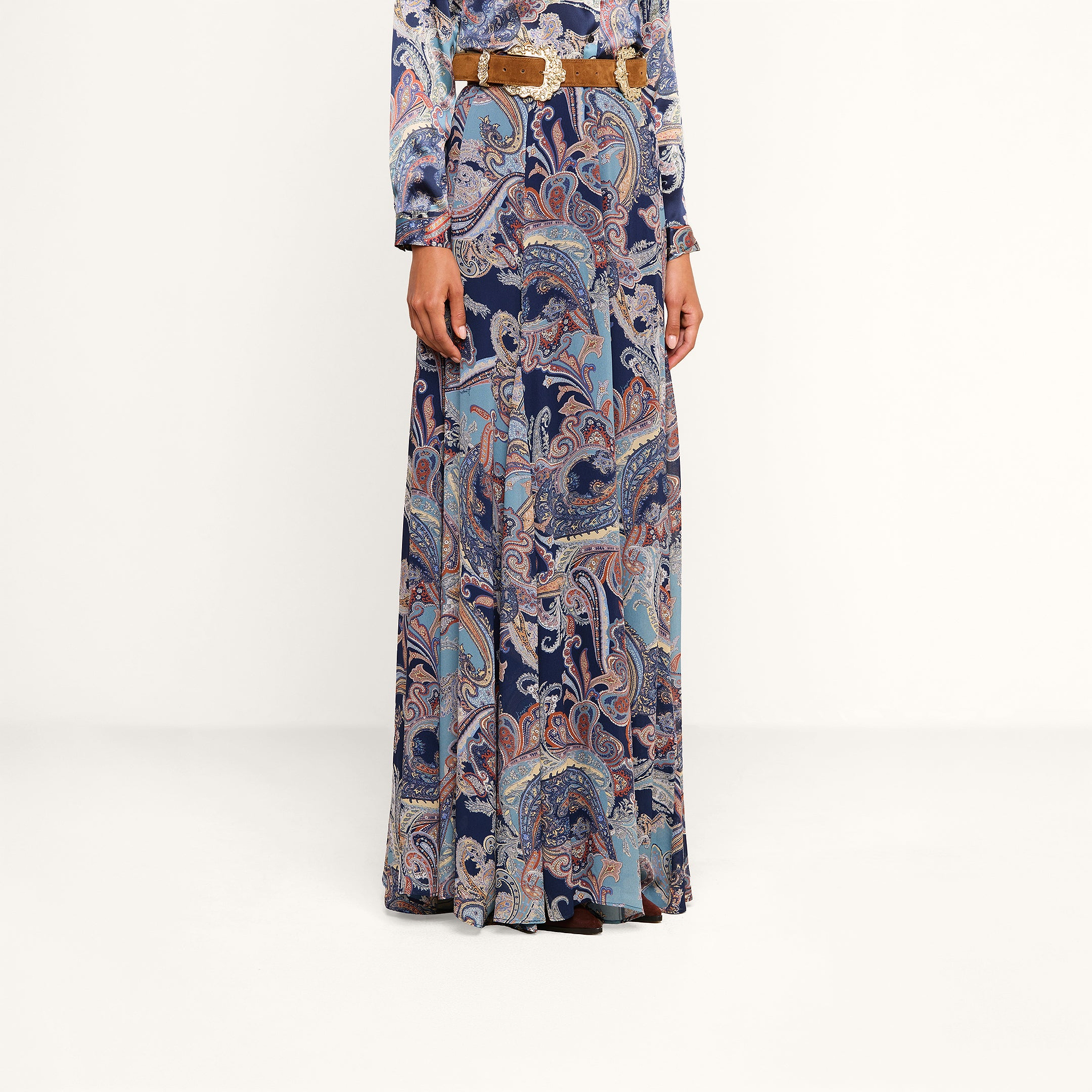 A person is wearing the Patrica Maxi Skirt, a floor-length paisley print skirt in shades of blue, purple, and pink, paired with a long-sleeved top and cinched at the waist with a brown belt. Their head is not visible in the image.