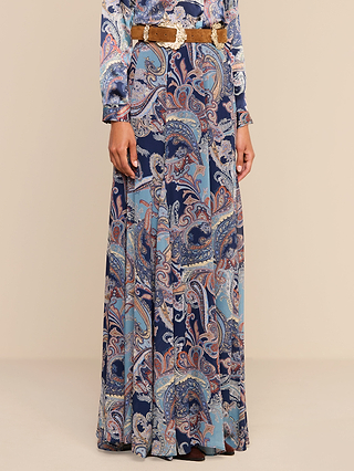 A woman models the Patrica Maxi Skirt, a long paisley-print piece in midnight blue and purple, styled with a wide tan belt and matching top. She stands with her hands relaxed by her sides against a plain beige background.