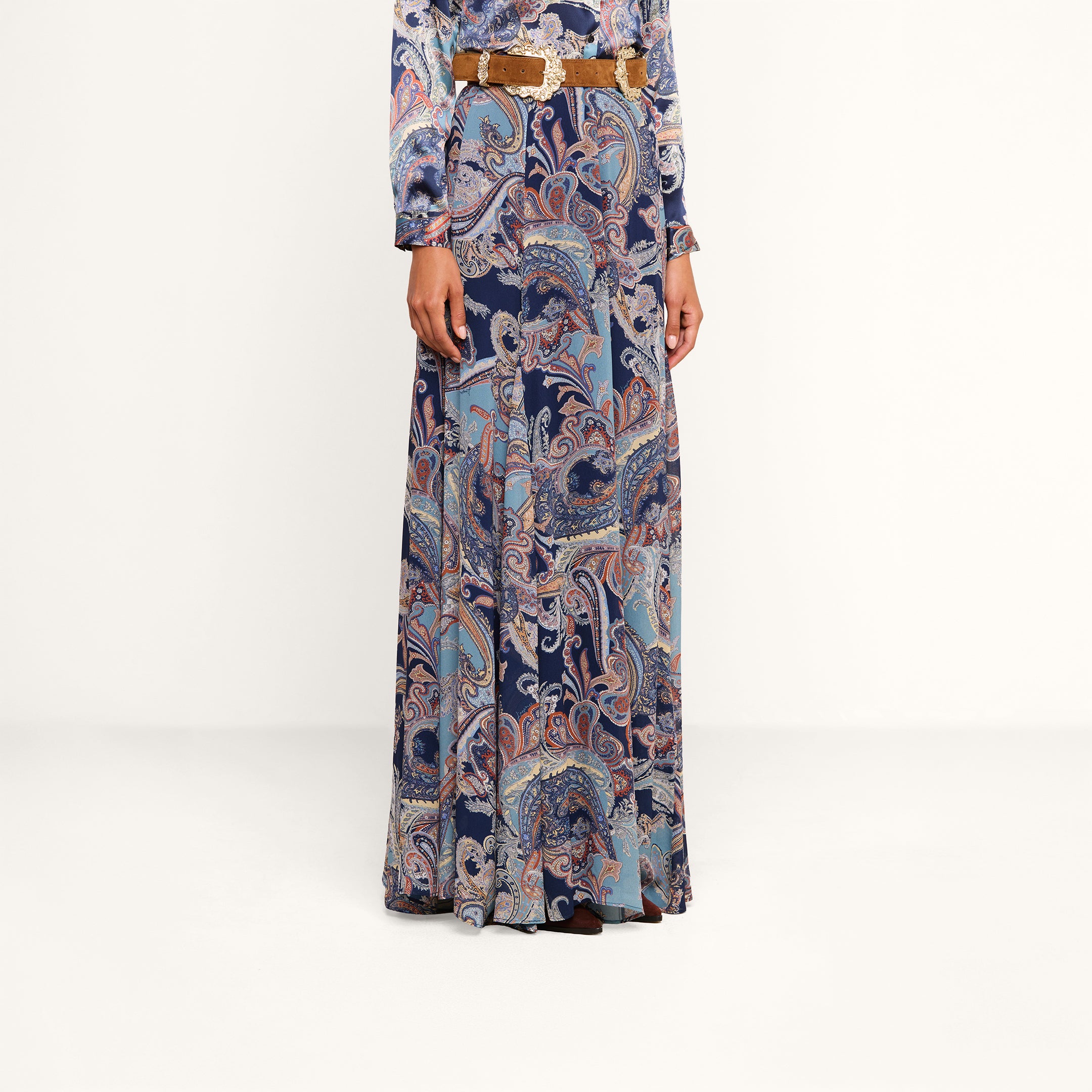 A person is wearing the Patrica Maxi Skirt, a floor-length paisley print skirt in shades of blue, purple, and pink, paired with a long-sleeved top and cinched at the waist with a brown belt. Their head is not visible in the image.