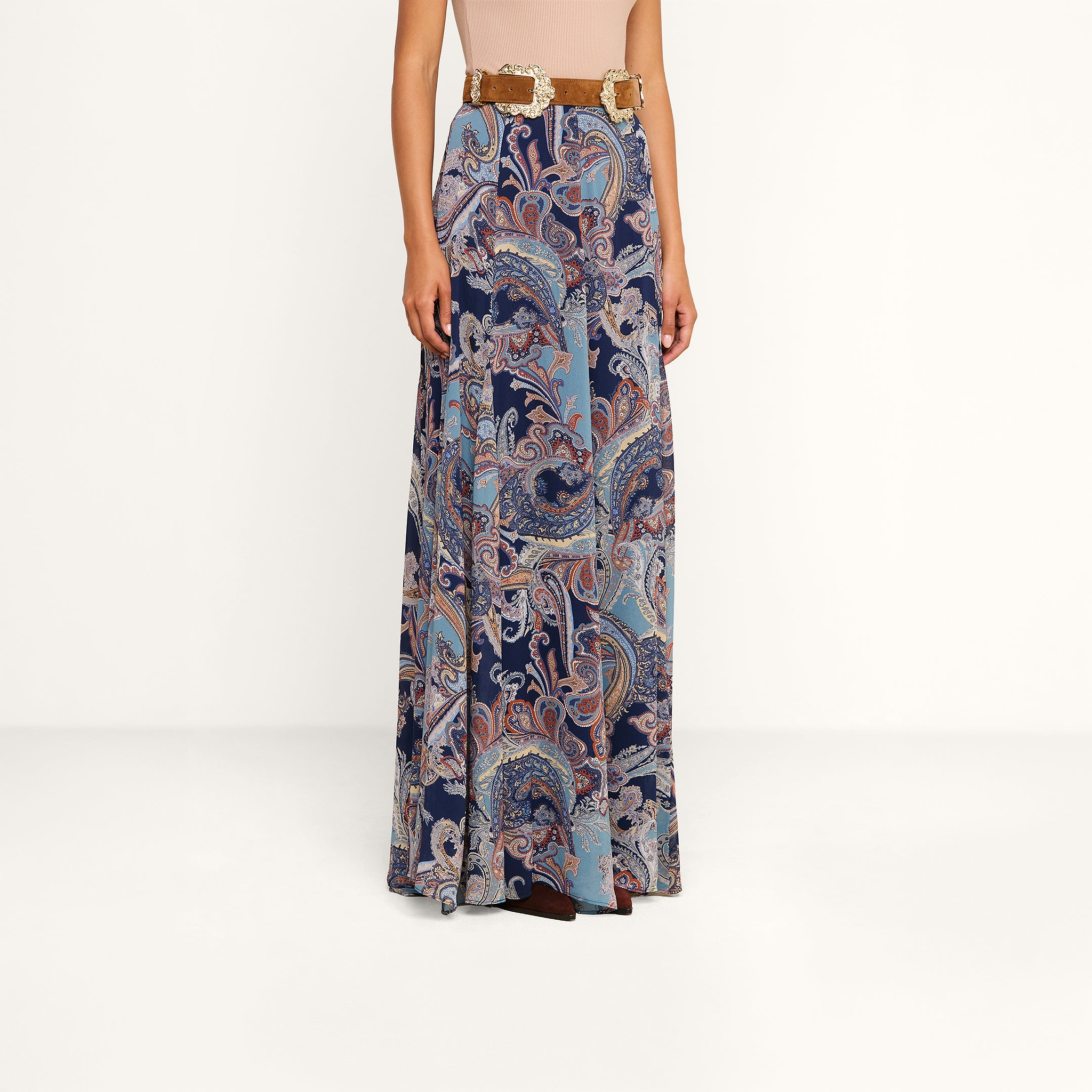 Wearing the Patrica Maxi Skirt, which features a long, flowing silhouette and a midnight blue and light brown paisley print, paired with a tan belt. The upper body and face are not visible against the plain white background.