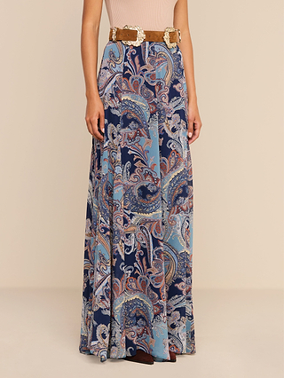 Wearing the Patrica Maxi Skirt in a midnight blue paisley print, paired with a light pink top and a tan belt featuring a large buckle, the person stands against a plain background.