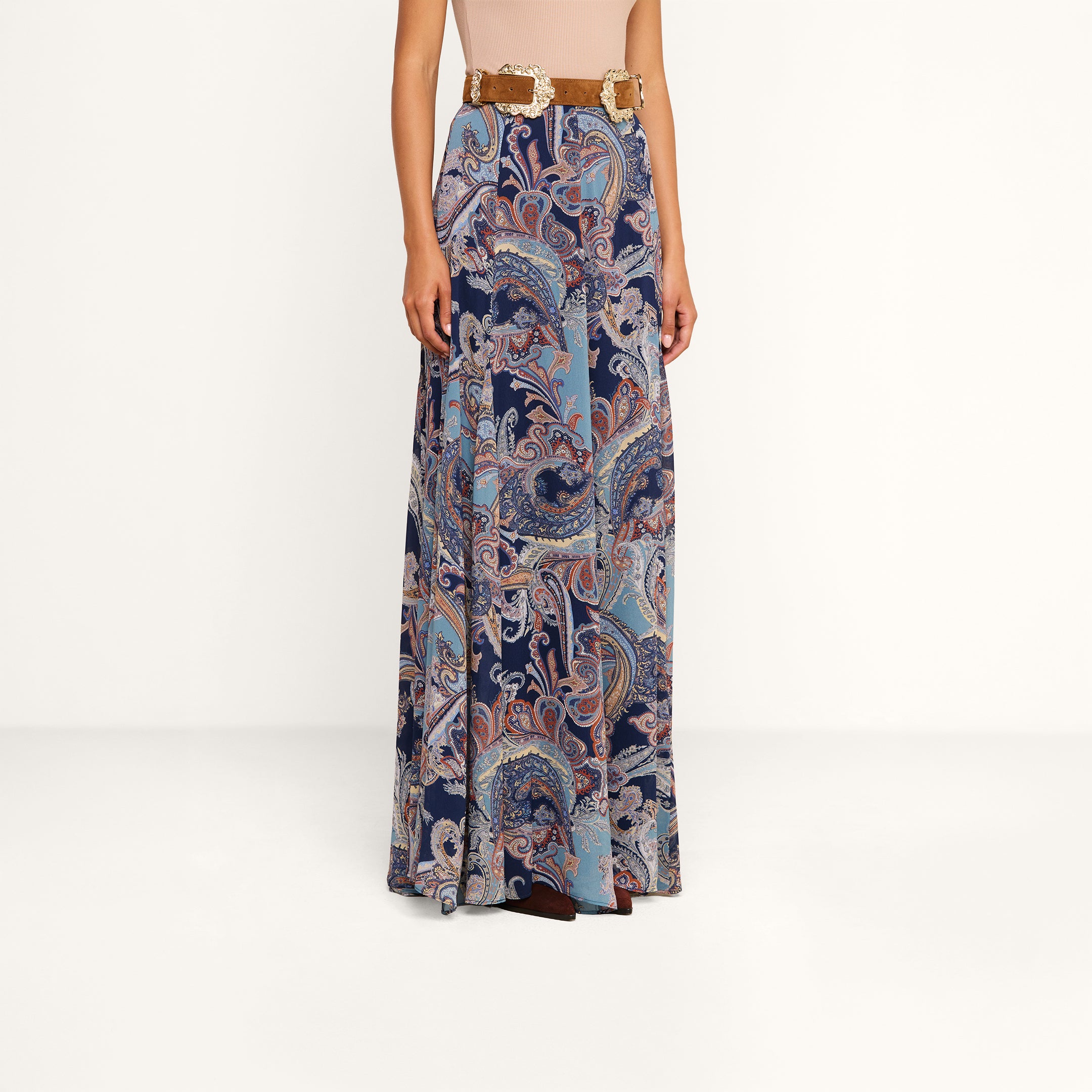 Wearing the Patrica Maxi Skirt, which features a long, flowing silhouette and a midnight blue and light brown paisley print, paired with a tan belt. The upper body and face are not visible against the plain white background.