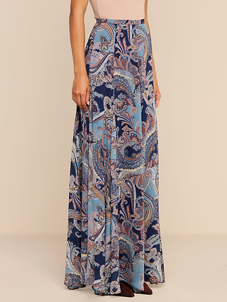 A woman wearing the Patrica Maxi Skirt – a high-waisted, midnight blue paisley print maxi skirt – paired with a blush pink sleeveless top and brown pointed-toe shoes, stands against a beige background.