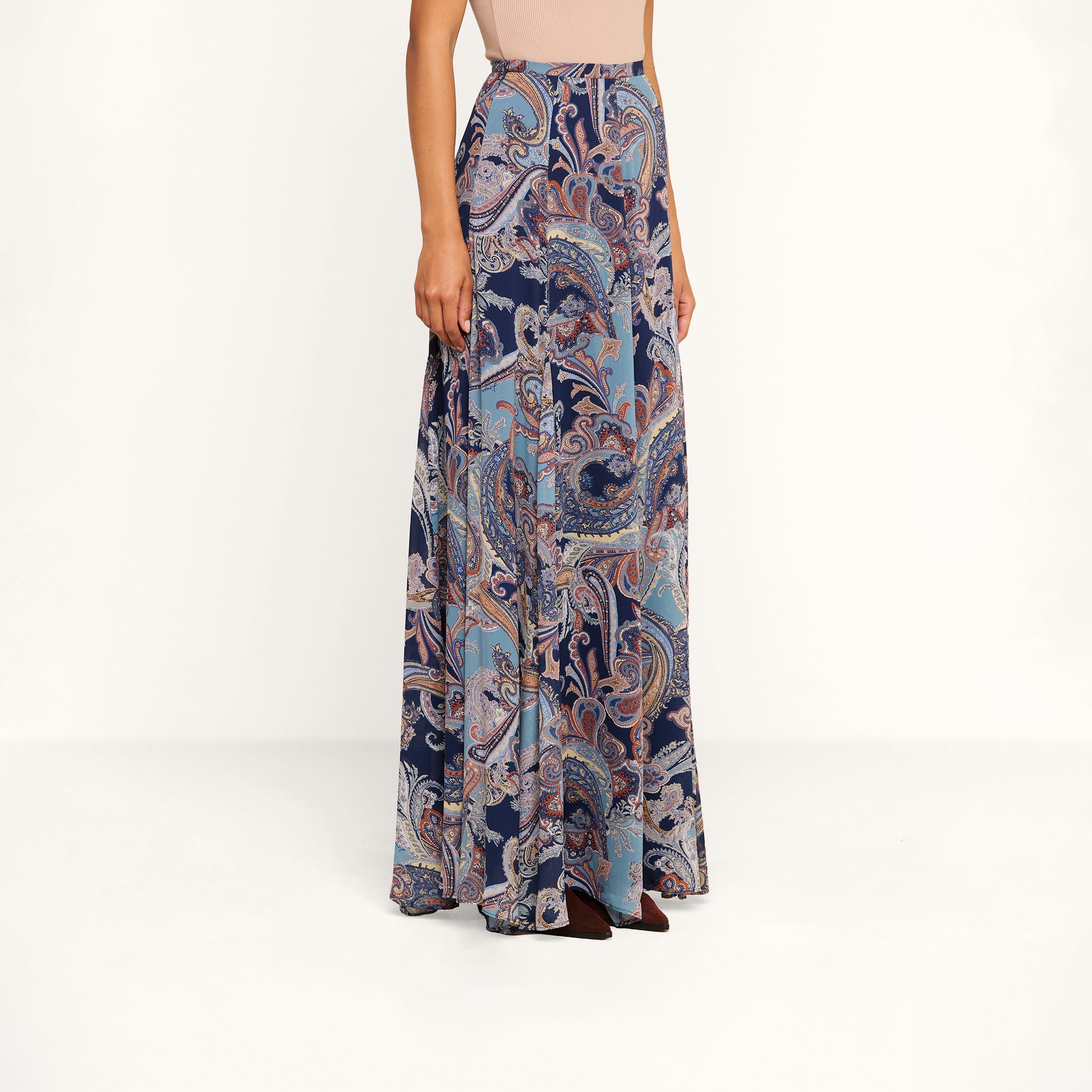 A woman wearing a sleeveless beige top paired with the Patrica Maxi Skirt—a high-waisted, floor-length skirt showcasing an intricate midnight blue and pink paisley print—stands against a plain white background.