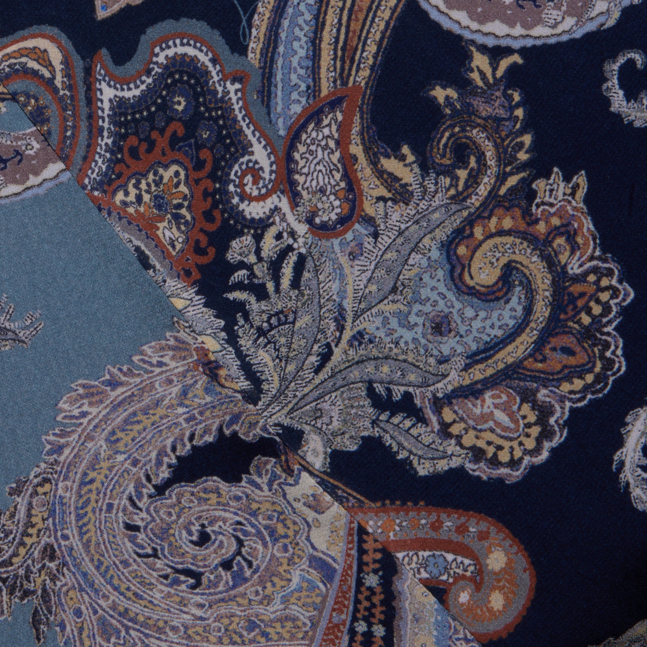 Close-up of overlapping fabric swatches from the Patrica Maxi Skirt, showcasing an intricate paisley print in blue, midnight blue, tan, and orange tones. The detailed designs feature swirling shapes and floral motifs.