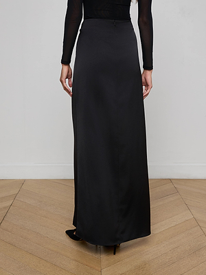 A person is standing indoors on a wooden floor, viewed from behind, wearing the L’AGENCE Emmarie Maxi Wrap Skirt in black with a high waist and a black long-sleeve top.