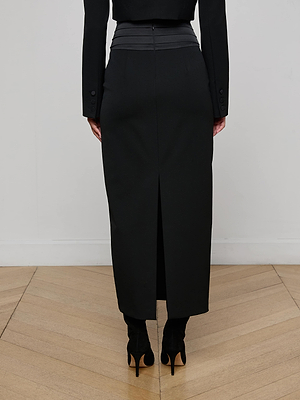 A person stands on a wooden floor, seen from the back, wearing the L'AGENCE Honesty Maxi Skirt with a satin waistband and back slit, styled with a black jacket and high heels for an elegant evening look.
