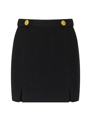 The Nelina Tweed Mini Skirt is a black mini skirt featuring gold-tone buttons on the waistband and small front slits near the hem, shown against a white background.