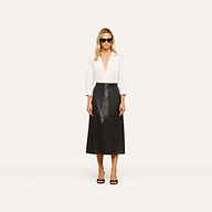 A woman stands facing forward against a plain light background, wearing sunglasses, a white long-sleeve blouse, black open-toe heels, and the Robyn Vegan Leather Skirt.