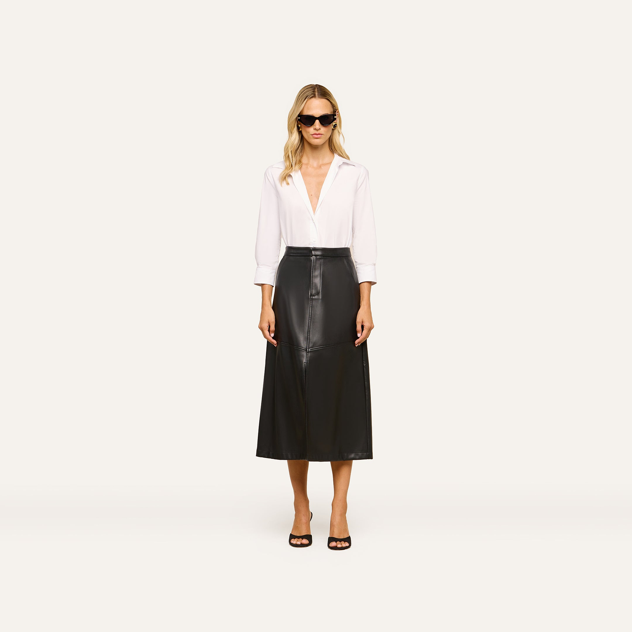 A woman stands facing forward against a plain light background, wearing sunglasses, a white long-sleeve blouse, black open-toe heels, and the Robyn Vegan Leather Skirt.