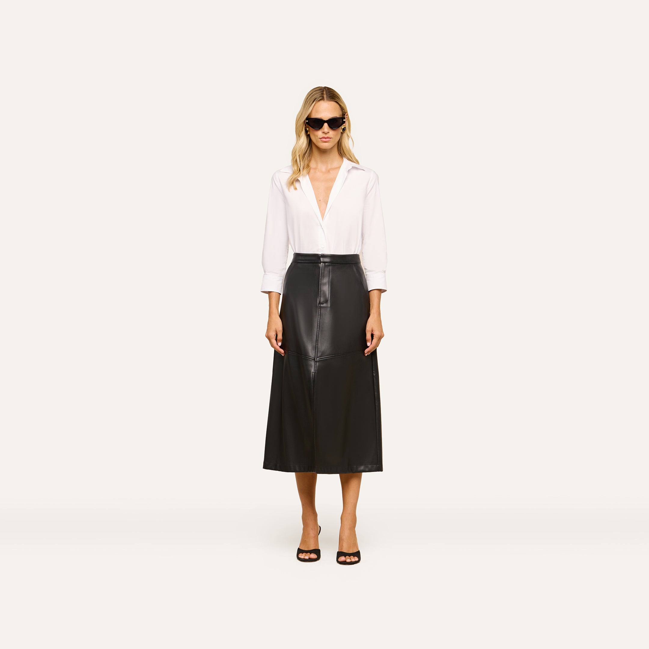 A woman stands facing forward against a plain light background, wearing sunglasses, a white long-sleeve blouse, black open-toe heels, and the Robyn Vegan Leather Skirt.