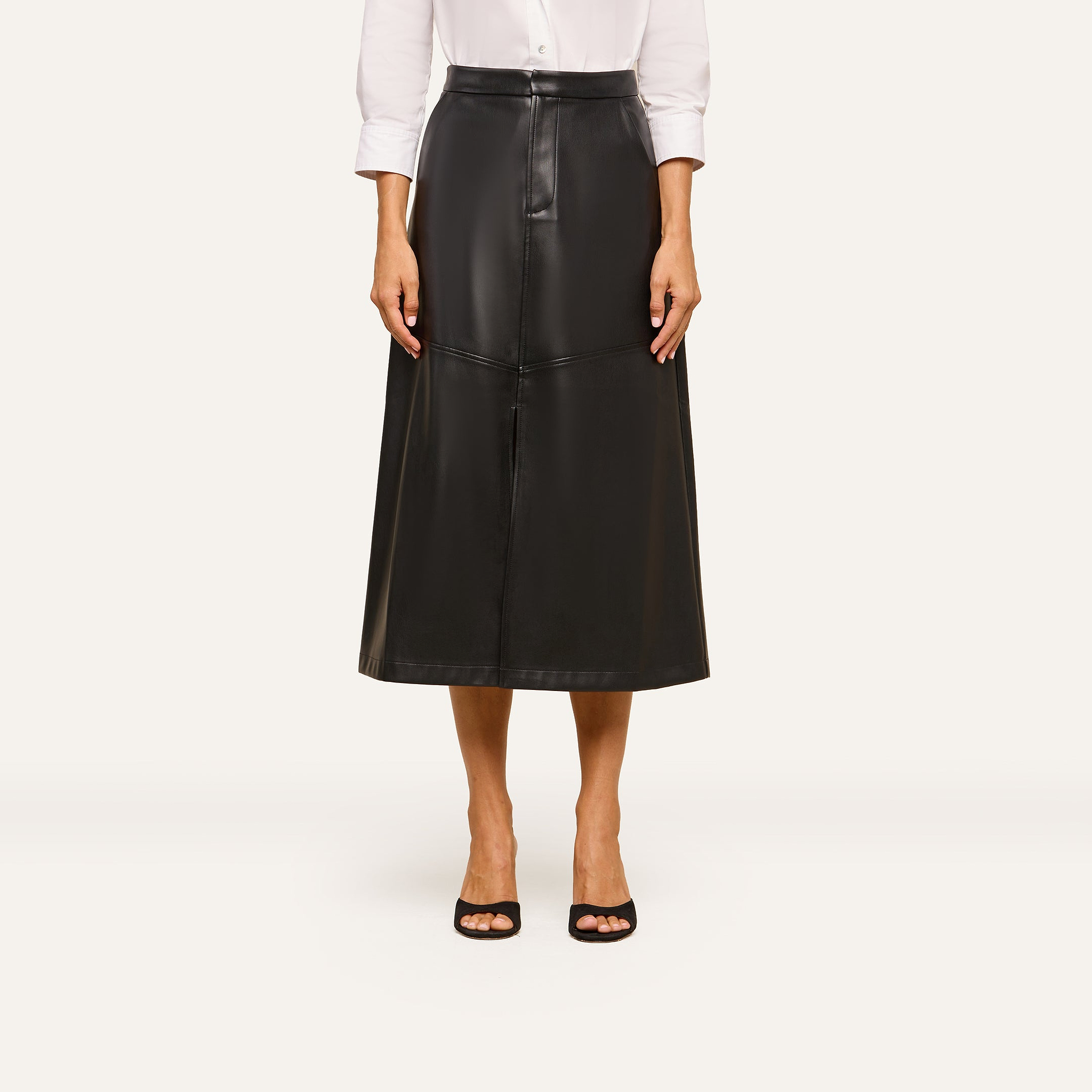 A person wears a white long-sleeve shirt tucked into the high-waisted Robyn Vegan Leather Skirt in black, featuring front seams, paired with black open-toe heels, and stands against a plain background.