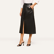 A woman wearing the Robyn Vegan Leather Skirt in black—a midi skirt with a front slit—paired with a white long-sleeve shirt and black high-heeled sandals. The photo captures her from the shoulders down against a light background.