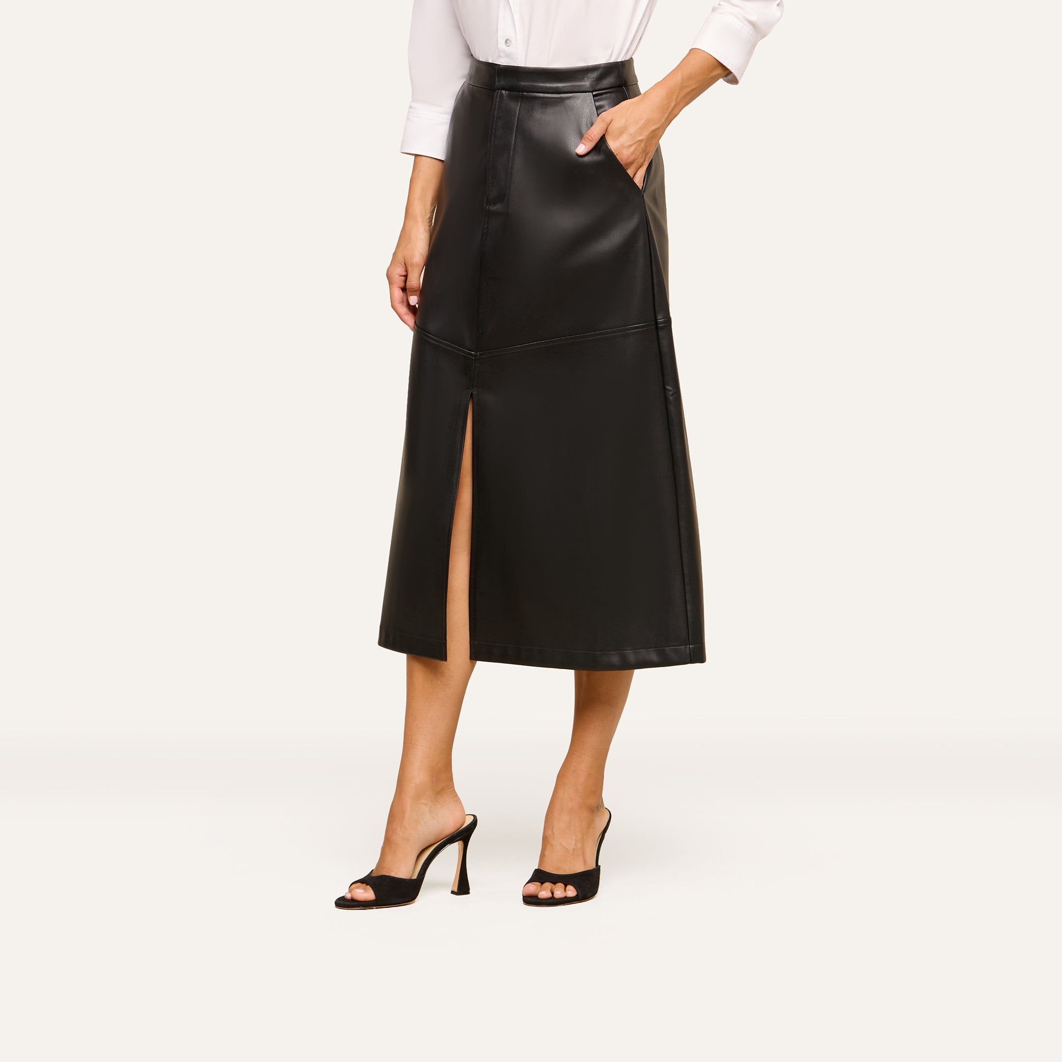 A woman wearing the Robyn Vegan Leather Skirt in black—a midi skirt with a front slit—paired with a white long-sleeve shirt and black high-heeled sandals. The photo captures her from the shoulders down against a light background.