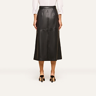 A person shown from the back stands in black heeled shoes against a plain background, wearing a white long-sleeve shirt and the Robyn Vegan Leather Skirt, a black A-line midi skirt.