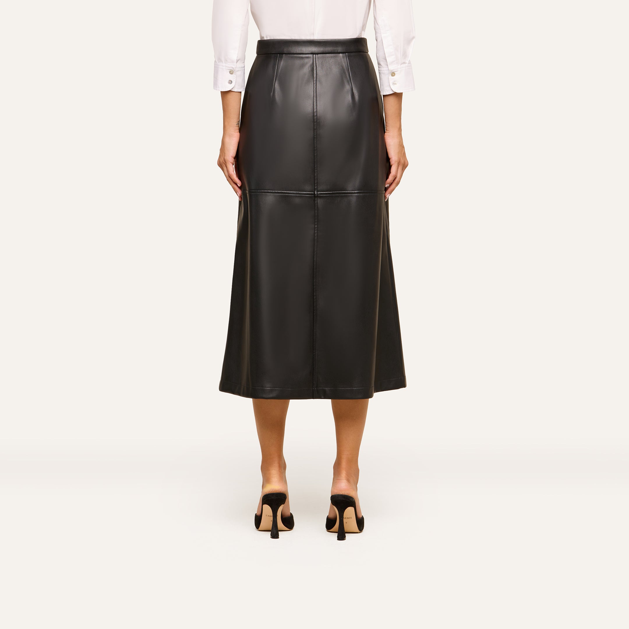 A person shown from the back stands in black heeled shoes against a plain background, wearing a white long-sleeve shirt and the Robyn Vegan Leather Skirt, a black A-line midi skirt.
