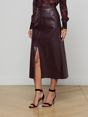 A woman stands indoors wearing the L'AGENCE Robyn Vegan Leather Midi Skirt in dark burgundy, featuring a high waist and front slit, paired with a sheer lace top and black heels. Her head is not visible in the image.