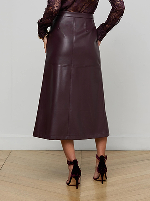 A person wearing the L'AGENCE Robyn Vegan Leather Midi Skirt in dark brown and high-heeled, open-back burgundy shoes stands on a wooden floor, seen from behind.