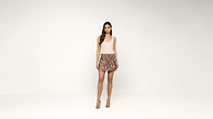 A woman stands against a plain white background, wearing a sleeveless beige top, a patterned mini skirt, and high-heeled sandals. She has long, loose hair and a neutral expression.