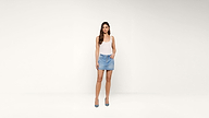 A woman stands against a plain white background, wearing a white tank top, a light blue denim mini skirt, and blue high heels. She has long, straight hair and one hand in her skirt pocket.