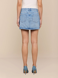 La modella di spalle indossa la minigonna Solene Jean, realizzata in denim 100 cotone con bottoni fantasia, abbinata a scarpe con tacco alto blu su uno sfondo beige.