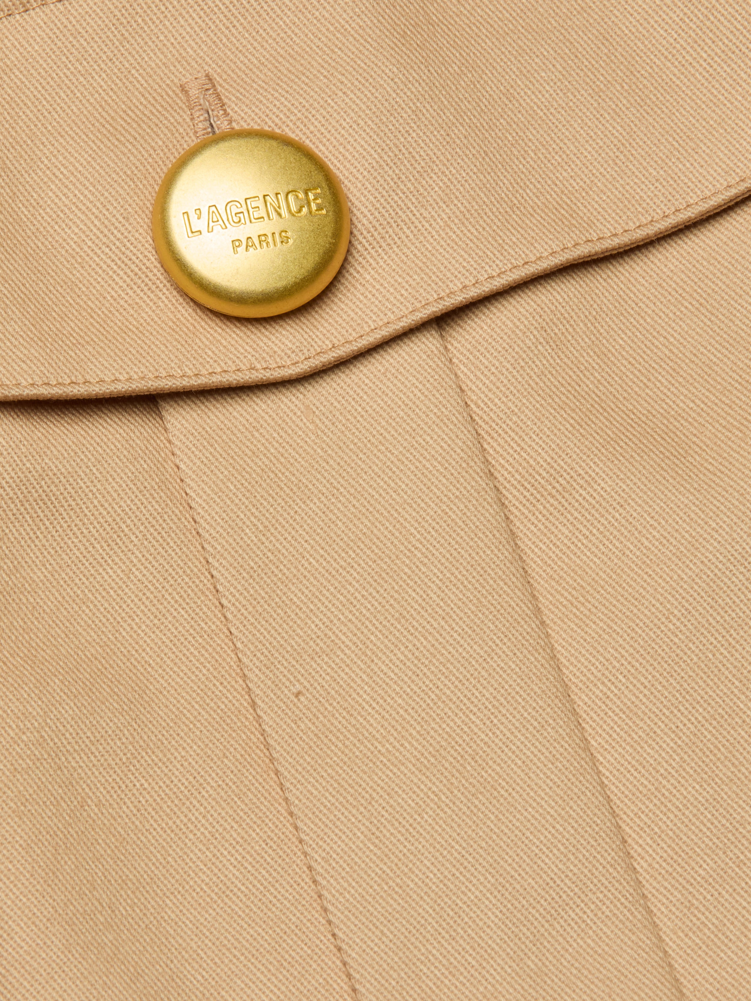 Close-up of the Maelis Mini Skirt in beige, featuring a gold button engraved with LAGENCE PARIS near the seam and flap, highlighting the tailored detail of this high-rise waist design.