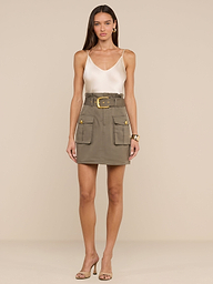 Una donna su uno sfondo beige indossa un top senza maniche in raso bianco, orecchini dorati, un braccialetto, sandali con tacco alto beige e la minigonna Maelis verde oliva con ampie tasche cargo e cintura larga.