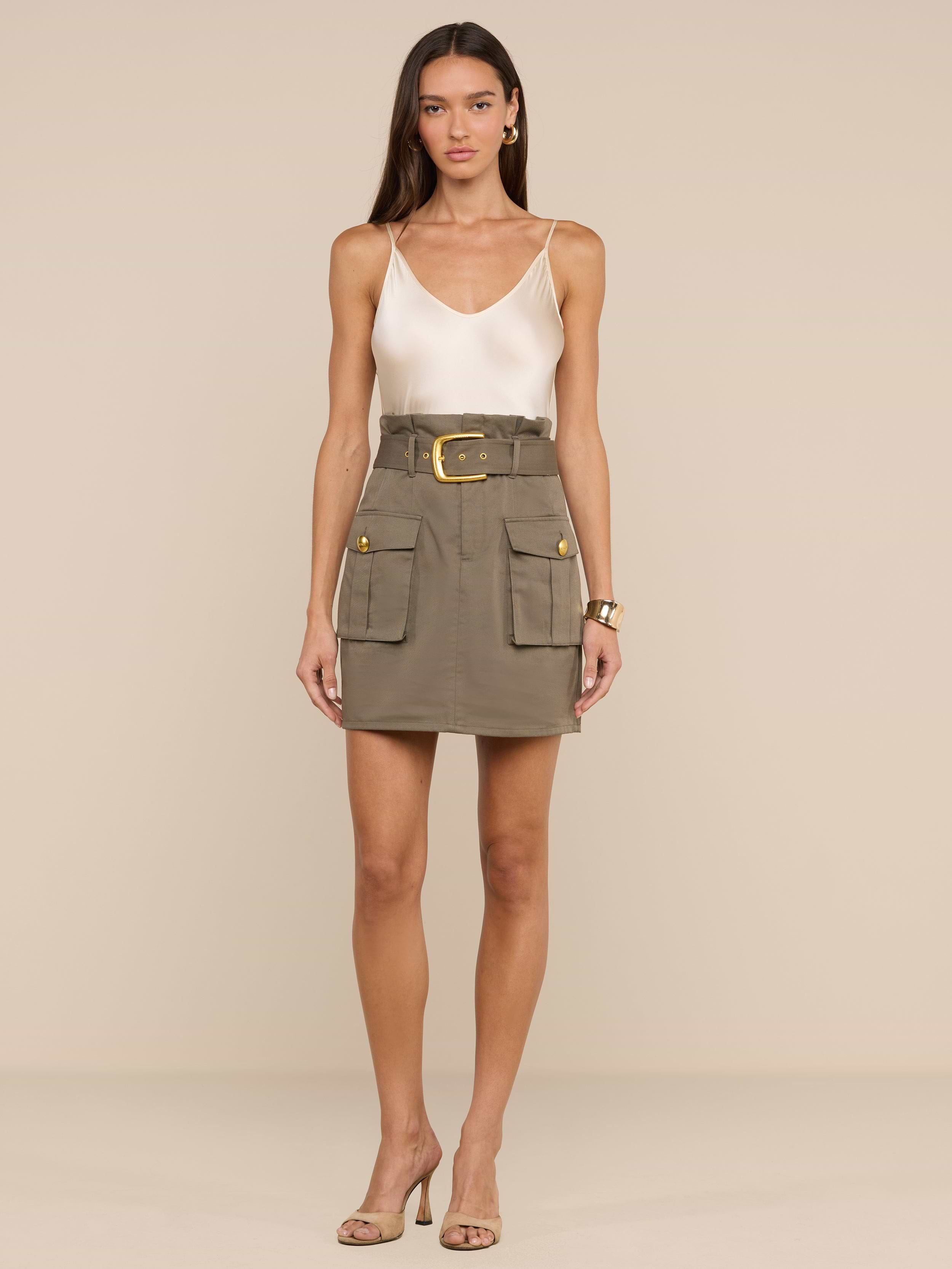 Una donna su uno sfondo beige indossa un top senza maniche in raso bianco, orecchini dorati, un braccialetto, sandali con tacco alto beige e la minigonna Maelis verde oliva con ampie tasche cargo e cintura larga.