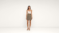 A woman stands in a minimal studio wearing a white tank top, olive green belted cargo skirt with pockets, gold jewelry, and heeled sandals. The background is plain white.